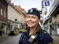 En ung poliskvinna ler mot kameran på en gata i Sverige. Hon bär uniform och utrustning, redo att rycka ut om larmet går.