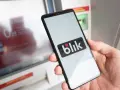 Dłoń trzyma smartfon z logo BLIK przed bankomatem.