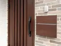 Puerta de madera oscura con tirador negro, buzón de madera y un timbre blanco. Varios tipos de timbre se pueden ver en la pared de ladrillos.
