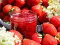 En burk med hemgjord jordgubbssylt omgiven av färska jordgubbar och blommor. Perfekt för ett enkelt jordgubbssylt recept.