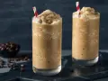 Dos vasos altos con granizado de café, decorados con pajitas rojas y blancas. Un tazón de granos de café en el fondo.