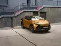 Złoty Toyota plug-in z pomarańczowym kablem ładowania podłączonym do stacji. Nowoczesna architektura w tle.