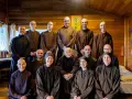 A group of monks and nuns, including an elder monk person in the center, pose for a photo in a room with wooden walls.