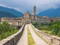 Ponte medievale in pietra che conduce a un borgo con chiesa e campanile, incastonato tra le verdi montagne degli Appennini Emilia Romagna.