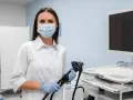Lekarka w masce i rękawiczkach trzyma endoskop, przygotowując się do gastroskopii. Obok monitor i sprzęt medyczny.