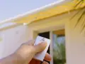 Main tenant une télécommande pour le réglage store banne manuel. Le store jaune est déployé sur une maison blanche.