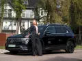 Kobieta w czarnym garniturze opiera się o czarnego Mercedesa EQB na tle białego domu.