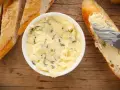 Knoblauchbutter selber machen ist einfach! Hier ist eine Schüssel mit selbstgemachter Knoblauchbutter, daneben Baguettescheiben und ein Messer, das Butter auf ein Brot streicht.