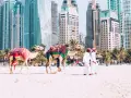 Dwa wielbłądy na plaży w Dubaju, obok palm i drapaczy chmur. Zastanawiasz się, ile się leci do Dubaju?