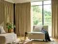Salon moderne avec canapé beige, fauteuil, plante verte et rideaux au **type de pli rideau** qui habillent une grande baie vitrée.