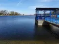 Spokojne jezioro średzkie z błękitną budowlą i pomostem. W tle drzewa i zabudowania.