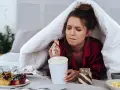 Ragazza sotto le coperte mangia gelato, dolci e pasta. La sua espressione suggerisce una forte fame pre ciclo.