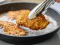 Une escalope de dinde panée aux graines de sésame est retournée dans une poêle avec de l'huile chaude.