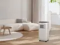 Un aire acondicionado portátil con tubo de escape conectado a la ventana, parte de unas modernas instalaciones hvac, en un salón con sofá beige.