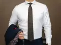 Elegancki mężczyzna w białej koszuli i modnym krawacie męskim w kropki, trzymający granatową marynarkę z wzorzystą podszewką.