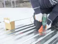 Pose sur une toiture bac acier isolé, un ouvrier visse une plaque avec une perceuse. Une boîte de vis est à proximité.