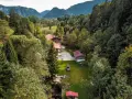 Idyllisches **Camping in den Bergen**: Ein Wohnmobil steht auf einer grünen Wiese neben einem Bach, umgeben von dichten Wäldern und traditionellen Holzhäusern.