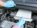 Ręce w niebieskich rękawiczkach wyjmują nowy filtr kabinowy z komory silnika. Wymiana filtra kabinowego to prosty sposób na czyste powietrze w aucie.