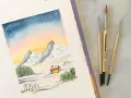 Une aquarelle montagne facile dépeint un paysage hivernal doux avec des montagnes enneigées, des chalets chaleureux et un ciel étoilé. Trois pinceaux sont posés à côté.