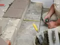 Mężczyzna mierzy taśmą, przygotowując się do tego, jak układać płytki na schodach zewnętrznych. Widać kielnie i wiadro z zaprawą.