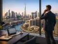 Biznesmen rozmawia przez telefon, patrząc na panoramę Dubaju. Na biurku laptop, kawa i notatnik. To symbol nowoczesnej pracy w Dubaju.