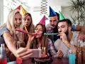 Radosne urodziny przyjaciółki! Grupa przyjaciół w kolorowych czapkach świętuje, dmucha w trąbki i robi selfie przy torcie.