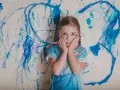 Dziewczynka z pędzlem w ręku, z dłońmi na policzkach, na tle niebieskich mazów. Czy asperger to choroba psychiczna?