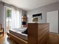 Un moderno **letto in mezzo alla stanza**, con un copriletto a motivi floreali, accanto a una poltrona leopardata e una cassettiera in legno.