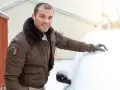 Mężczyzna w kurtce z futrzanym kołnierzem, uosabiający modę skandynawską, czyści samochód ze śniegu.