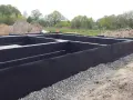Fundament domu w trakcie budowy, z czarną membraną i otoczakami. Trwa ocieplenie fundamentów, przygotowanie do dalszych prac.