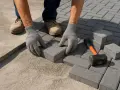 Mani guantate posano blocchi per creare un pavimento in cemento per esterno. Martello e mattoni a portata di mano.