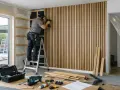 Man på en stege, som använder en spikpistol för att montera akustikpanel på en vägg. Verktyg och material ligger utspridda på golvet.