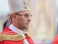 Biskup w stroju pontyfikalnym, z mitrą na głowie, w okularach. W tle widać fragment obrazu.