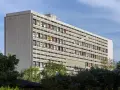 Długi budynek mieszkalny w stylu modernizm, z rzędami balkonów w kolorowych obramowaniach, otoczony zielenią.