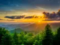 Sunset over rolling blue mountains with lush green trees in the foreground.
