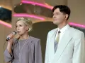 Kto prowadził czar par? Ona z mikrofonem, on w jasnej marynarce, na tle neonowych świateł.