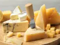 Różnorodne rodzaje serów żółtych, w tym pleśniowe i twarde, ułożone na drewnianej desce. Nóż do sera wbity w kawałek parmezanu.