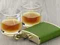 Zwei Gläser mit bernsteinfarbenem Whiskey und ein grüner Taschenflachmann. Ein 4 cl Whiskey hat ca. 90 Kalorien.
