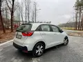 Biały Honda Fit stoi na mokrej, brukowanej nawierzchni. W tle drzewa i budynki.
