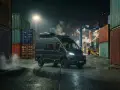 Mörk Fiat Ducato står vid en hamn, omgiven av containrar. Rök stiger från marken, kanske ett tecken på fiat ducato automatlåda problem.