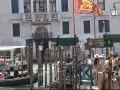 Un gruppo di innamorati a Venezia attende una gondola, con un vaporetto sullo sfondo e il leone di San Marco che sventola.