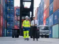Dwóch mężczyzn w kaskach na tle kontenerów. Jeden w odblaskowym stroju roboczym, drugi z tabletem. Spedycja transport krajowy w akcji.