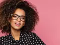 Une jeune femme aux cheveux bouclés porte un maquillage éclatant et des lunettes noires, souriant sur fond rose.
