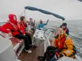Grupa osób na jachcie, w kamizelkach ratunkowych, cieszy się rejsem. Jedna z osób trzyma ster, inni obserwują morze. Wszyscy wyglądają na zadowolonych, jakby właśnie zdobyli patent żeglarski.