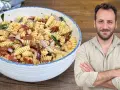 Un uomo sorride accanto a una ciotola di pasta fredda con ceci, pomodori secchi e cipolla rossa.