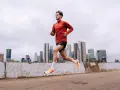 Mężczyzna w czerwonej bluzie i czarnych spodenkach biegnie, trenując jak przebiec 10 km. W tle miejski krajobraz z drapaczami chmur.