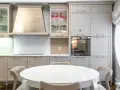 Cocina moderna con mesa redonda blanca y sillas. Gabinetes grises y beige, campana extractora de acero inoxidable. Ideal para ver catálogos de cocinas y precios.