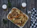 Pollo asado con patatas y zanahorias en una bandeja blanca, con cerveza y servilleta a cuadros.