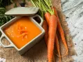 Un tazón de cremoso puré de zanahoria, adornado con hebras de zanahoria rallada, junto a zanahorias frescas y verdes.