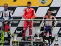 Podio con tre motociclisti famosi in tuta: Rossi, Stoner e Lorenzo. Vittoria celebrata con trofei.